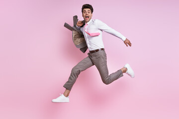 Happy man in shirt and tie carrying jacket on shoulder while jumping against pink background