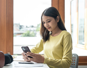 Young woman student in sweater is using smartphone to chatting with classmate while taking a break