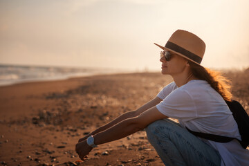 Young happy relaxed woman in a hat sitting on the seashore at sunset