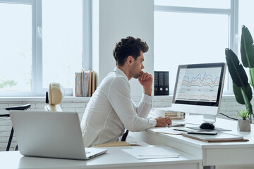 Thoughtful man in shirt and tie using computer while sitting at his working place in office