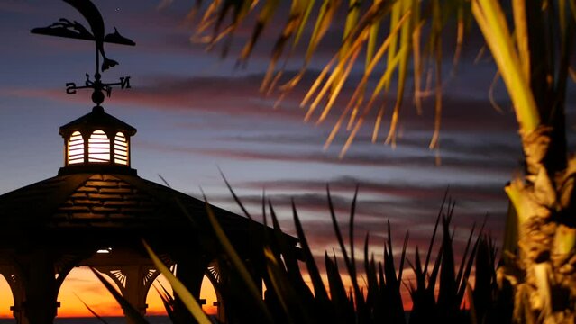 Sunset cloudscape and gazebo silhouette on beach, twilight dusk, clouds in dramatic purple pink sky, California coast. Wooden romantic cosy alcove and palm tree. Weather wind vane or roof weathercock.