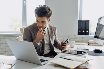 Concentrated young man using laptop and holding smart phone while sitting in office