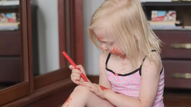 Little blonde girl sits on floor and enthusiastically paints arms and legs with red felt-tip pen. Happy childhood.