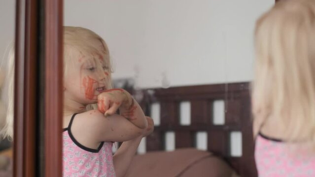 Little blonde girl spins around mirror and paints herself with red marker. Child fooling around.