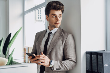 Fototapeta premium Handsome young man holding smart phone and looking through a window in office