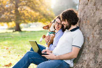 Happy freelancers and dog sit with laptop in autumn park under tree. People and puppy having fun together outdoors. Pet owners enjoying working, shopping online in nature. Authentic lifestyle moments - Powered by Adobe