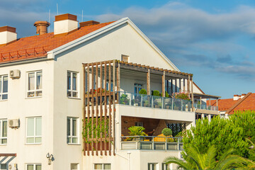 Fototapeta na wymiar Residential building with roof tiles and large terraces with wooden elements, glass balconies and decorative leafy flowering plants, sun loungers and chairs.