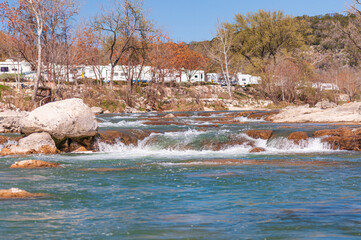 A winter day by the Guadalupe River, surrounded by rock and trees. A rapid flow of water rushes past a recreations vehicle along its shore.