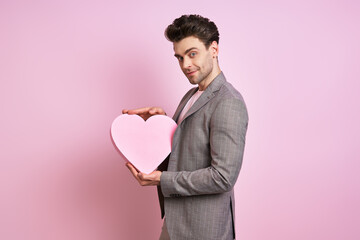 Happy young man in suit holding paper heart while standing against pink background