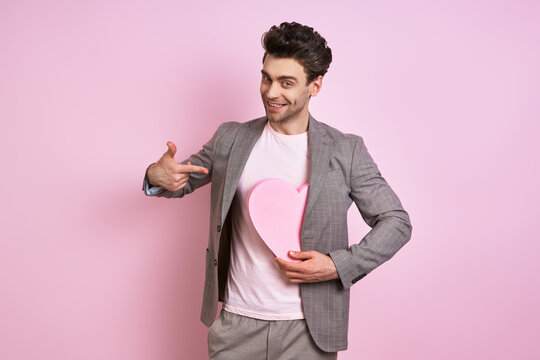 Happy young man in suit pointing paper heart while standing against pink background