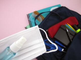      As COVID-19 prevention goes back to school and new normal concept. Top view of backpack with school supplies, mask and disinfectant gel on pink background.                              
