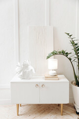 Stylish white bedside table with decor, lamp and white abstract gypsum painting  against a textured white wall. Potted plant at home. Nobody.