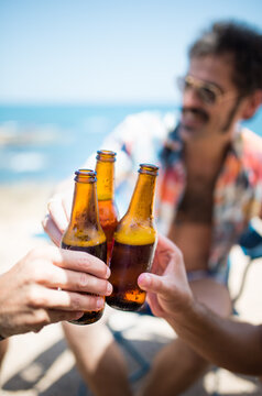 foto vertical con copyspace de grupo de amigos felices bebiendo y chocando botellines de cerveza fr&iacute;a en un camping al lado de la playa en verano