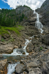 grosser Wasserfall der Engstligen in Adelboden. Das Wasser fett &uuml;ber eine hohe Felswand auf Ger&ouml;ll, Steine und Felsen. Am Ufer hat es Gras, Blumen und B&auml;ume