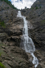 Obraz premium grosser Wasserfall der Engstligen in Adelboden. Das Wasser fett über eine hohe Felswand auf Geröll, Steine und Felsen. Am Ufer hat es Gras, Blumen und Bäume