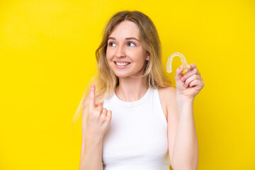 Blonde English young girl holding invisible braces isolated on yellow background intending to realizes the solution while lifting a finger up