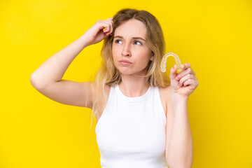 Blonde English young girl holding invisible braces isolated on yellow background having doubts and...