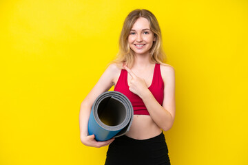 Young sport caucasian woman going to yoga classes while holding a mat isolated on yellow background pointing to the side to present a product