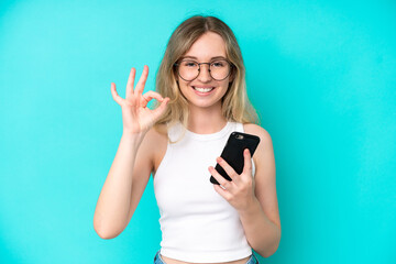 Blonde English young girl isolated on blue background using mobile phone and doing OK sign