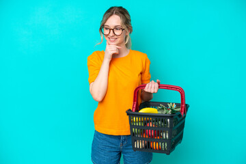 Blonde English young girl holding a shopping basket full of food isolated on blue background...