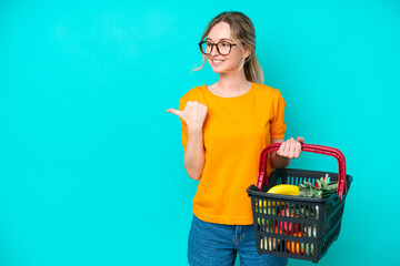 Blonde English young girl holding a shopping basket full of food isolated on blue background pointing to the side to present a product