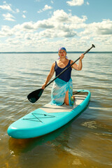 A Jewish woman in a pareo and a headdress stands on a SUP board with an oar on the lake.