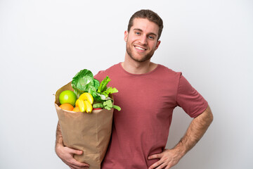 Young caucasian man holding a grocery shopping bag isolated on white background posing with arms at...