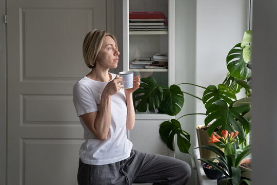 Tranquil middle-aged female enjoys hot drink from mug standing at windowsill. Mature blonde woman enjoys view outside window drinking hot coffee. Lady thinks about lifestyle and forthcoming day