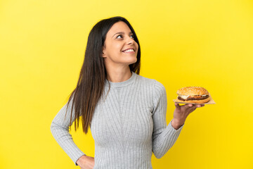 Young caucasian woman holding a burger isolated on yellow background thinking an idea while looking...