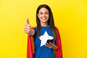 Super Hero woman isolated on yellow background using mobile phone while doing thumbs up