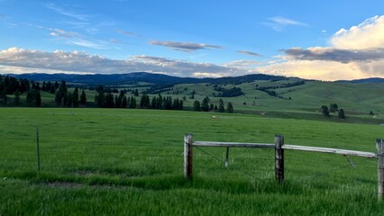 Wooden gate and barbed wire fencing in foreground, with grass land and pine-covered hills beyond. Overcast, dark, with a few clouds overhead. Montana, USA