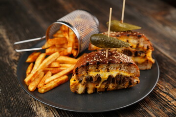 Delicious beef burger cut in half with cheese, pickled, tomatoes, mustard, and fries on a black plate on wooden background. Fast food.