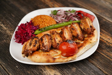 Grilled Turkish chicken wings served with pita, garnish,  onions, bulgur, roasted green pepper, and roasted tomato on white plate on wooden background. Served special restaurant presentation