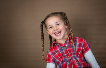 Adorable laughing toddler in braids and plaid shirt isolated on brown background - 521885315