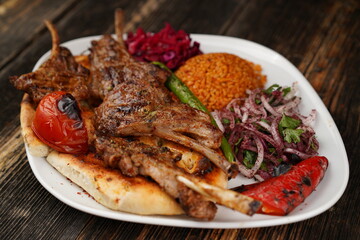 Grilled lamb chops on a white plate on wooden background. Grilled lamb with pita, garnish,  onions, bulgur, roasted green pepper, and roasted tomato. Turkish name: kuzu pirzola. 