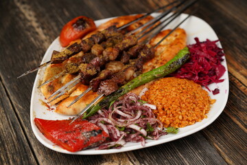 A skewered meat kebab with grilled tomato, onion, and rise. Grilled shish meat served with a white plate on wooden background. Traditional grilled kebabs.