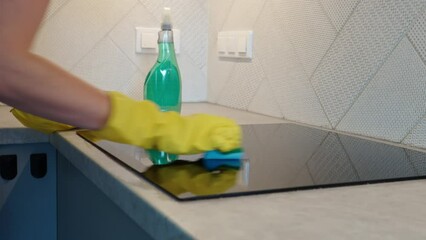 Woman in yellow rubber gloves cleans kitchen induction hob with cleaning spray and sponge. Clear kitchen appliance, Daily chores and housework