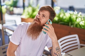 Young redhead man talking on the smartphone sitting on table at coffee shop terrace