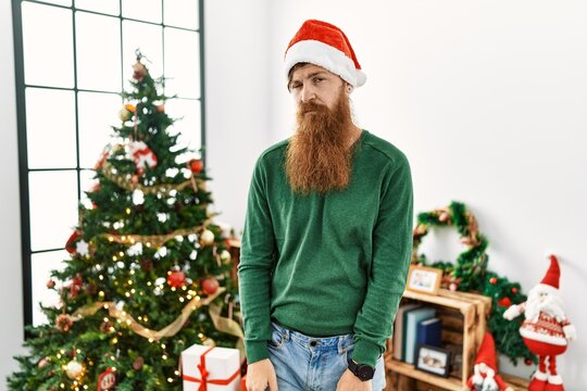 Redhead man with long beard wearing christmas hat by christmas tree looking sleepy and tired, exhausted for fatigue and hangover, lazy eyes in the morning.