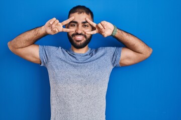 Middle east man with beard standing over blue background doing peace symbol with fingers over face, smiling cheerful showing victory