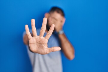 Middle east man with beard standing over blue background covering eyes with hands and doing stop...