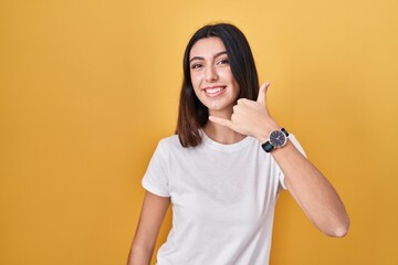 Young beautiful woman standing over yellow background smiling doing phone gesture with hand and fingers like talking on the telephone. communicating concepts.