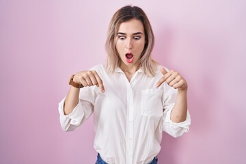 Young beautiful woman standing over pink background pointing down with fingers showing advertisement, surprised face and open mouth