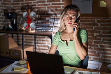 Young beautiful woman working at the office at night speaking on the phone puffing cheeks with funny face. mouth inflated with air, crazy expression.