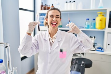 Young woman working at scientist laboratory holding blood sample smiling and laughing hard out loud...
