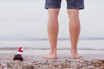 Young man standing on the sand by sea. Single bottle of whisky with Santa hat in sand of the sea, legs near of it. Christmas party, Xmas time, festive decoration, soft focus. Seasonal holidays concept