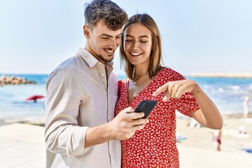 Young hispanic couple on vacation smiling happy using smartphone at the beach
