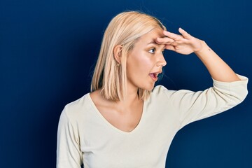 Beautiful blonde woman wearing casual sweater very happy and smiling looking far away with hand over head. searching concept.