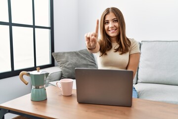 Fototapeta na wymiar Young brunette woman using laptop at home drinking a cup of coffee showing and pointing up with finger number one while smiling confident and happy.