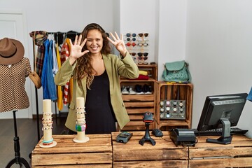 Beautiful hispanic woman working at fashion shop showing and pointing up with fingers number nine while smiling confident and happy.